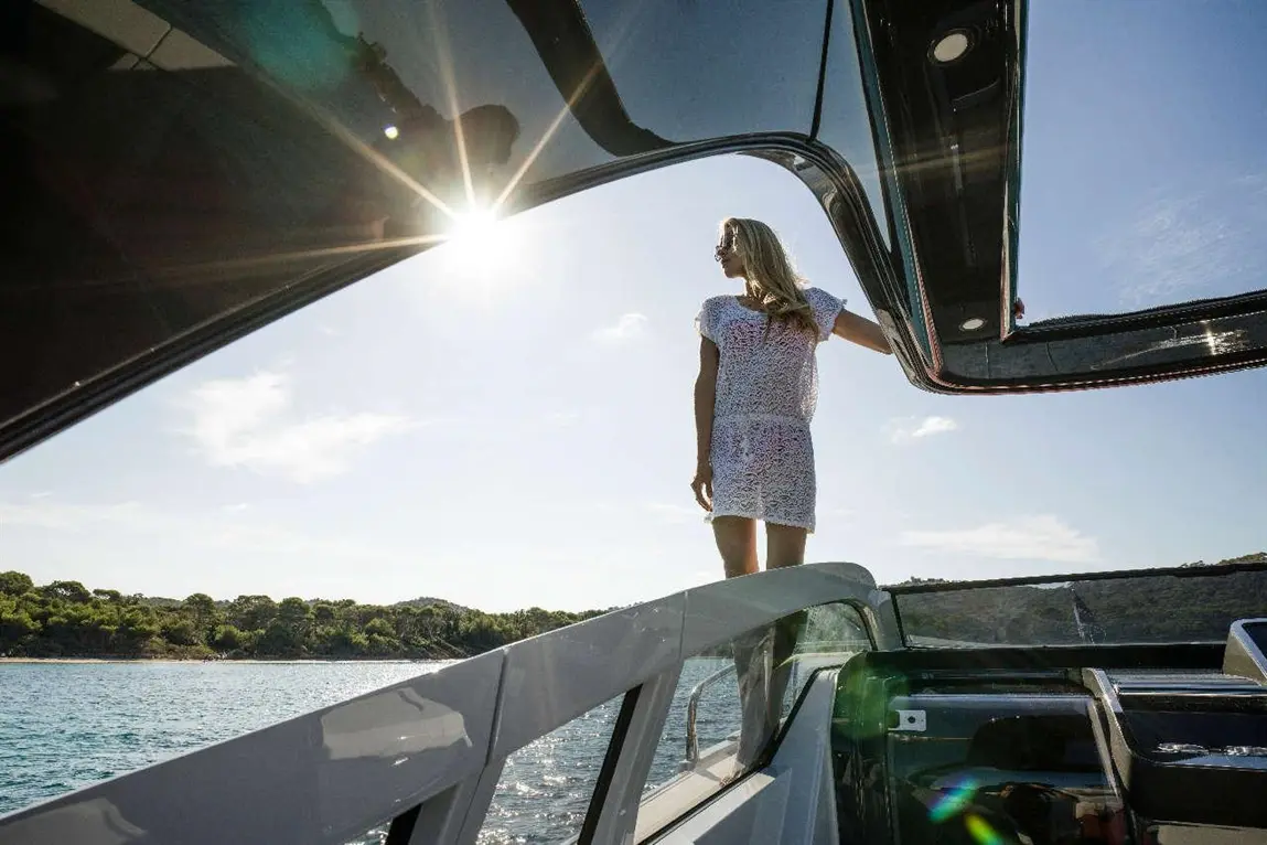 Woman in a white dress stands on a yacht, enjoying the sun and sea view, with lush greenery in the background.